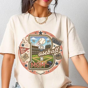 May include: Cream-colored t-shirt featuring a baseball-themed graphic. The design includes a large letter "D" with a baseball field, baseballs, and the words "Baseball Mom." Additional elements include a baseball glove, a helmet, and a home run flag.