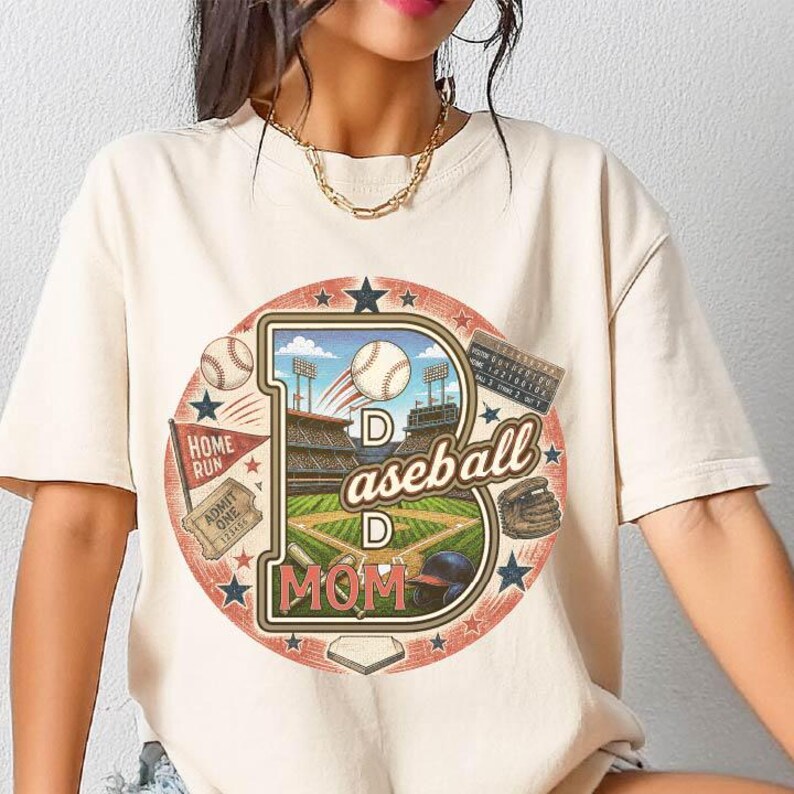 May include: Cream-colored t-shirt featuring a baseball-themed graphic. The design includes a large letter "D" with a baseball field, baseballs, and the words "Baseball Mom." Additional elements include a baseball glove, a helmet, and a home run flag.