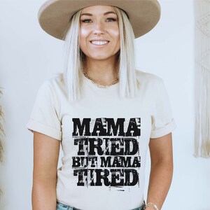 Pu&ograve; includere: Una maglietta beige con una grafica nera che recita "Mama Tried But Mama Tired".