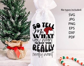 Puede incluir: Bolsa blanca de botella de vino de arpillera con el texto "SO TELL ME WHAT you want WHAT YOU REALLY really want" y un gráfico de gorro de Papá Noel. Un pequeño árbol de Navidad y una guirnalda de cuentas están en el fondo.