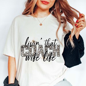 Peut inclure: T-shirt blanc avec le texte "livin' that COACH wife life" en lettres noires avec un motif l&eacute;opard.