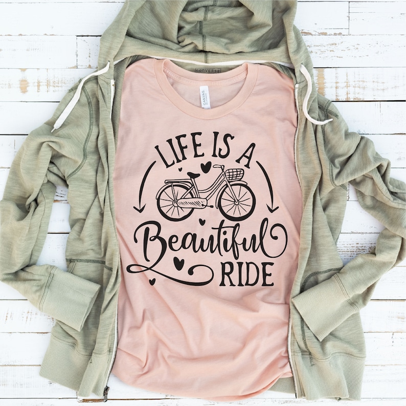 May include: A pink t-shirt with the text "Life is a Beautiful Ride" in black lettering. A black and white illustration of a bicycle with a basket is above the text. There are small hearts around the bicycle and text.