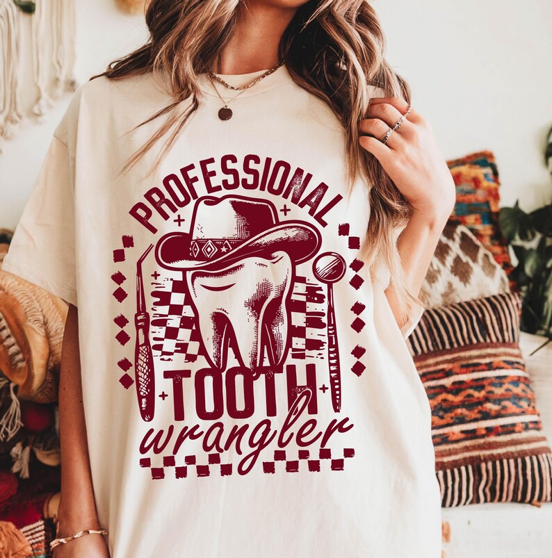 Op de afbeelding: Cr&egrave;mekleurig T-shirt met een bordeauxrode afbeelding. De afbeelding bevat de woorden "PROFESSIONAL TOOTH WRANGLER" rond een tand met een cowboyhoed, met tandheelkundige instrumenten. Het ontwerp heeft een dambordpatroon.