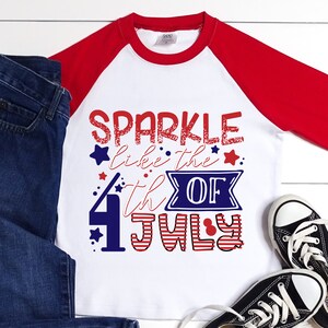 Puede incluir: Camiseta blanca y roja de manga raglan con un gráfico azul y rojo que dice "Sparkle like the 4th of July".