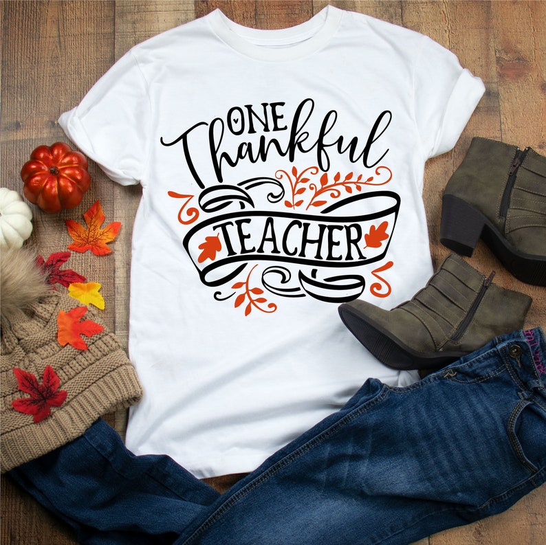 May include: White t-shirt with the text "One Thankful Teacher" in black with orange leaves and a black banner. The shirt is laying on a wooden surface with jeans, boots, and fall leaves.