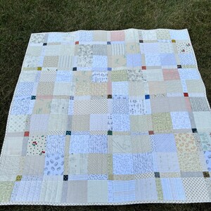 May include: A handmade patchwork quilt with a variety of fabric patterns in shades of white, beige, and pastel colors. The quilt is composed of numerous squares, some with floral and geometric designs, and is bordered with a dotted trim. The quilt is laid out on green grass.