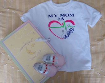 Può includere: Maglietta bianca con un cuore rosso e una linea ECG verde e rosa. Il testo "My Mom is a Nurse" è scritto all'interno del cuore. Una scarpa da bambino rosa è su un libro giallo con il testo "wonderful memories".