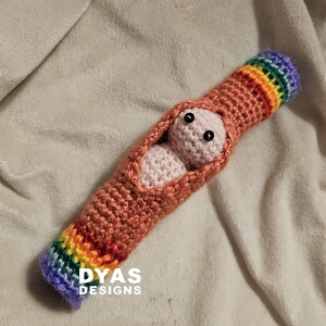May include: A handmade crochet baby cocoon in shades of orange and pink, with a rainbow-colored cuff. The cocoon holds a small, light pink crocheted baby with black eyes. The text "DYAS DESIGNS" is visible.