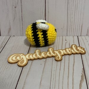 Puede incluir: Una abeja de ganchillo hecha a mano con rayas amarillas y negras, alas blancas y peque&ntilde;os ojos negros. La abeja est&aacute; sobre una superficie de madera. Un letrero de madera con el texto "@jadedpuris" est&aacute; en primer plano.