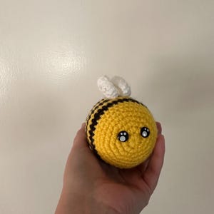 Puede incluir: Una abeja de ganchillo hecha a mano con cuerpo amarillo, rayas negras y alas blancas. La abeja tiene dos ojos negros y blancos, lo que le da una apariencia linda. La abeja se sostiene en una mano.