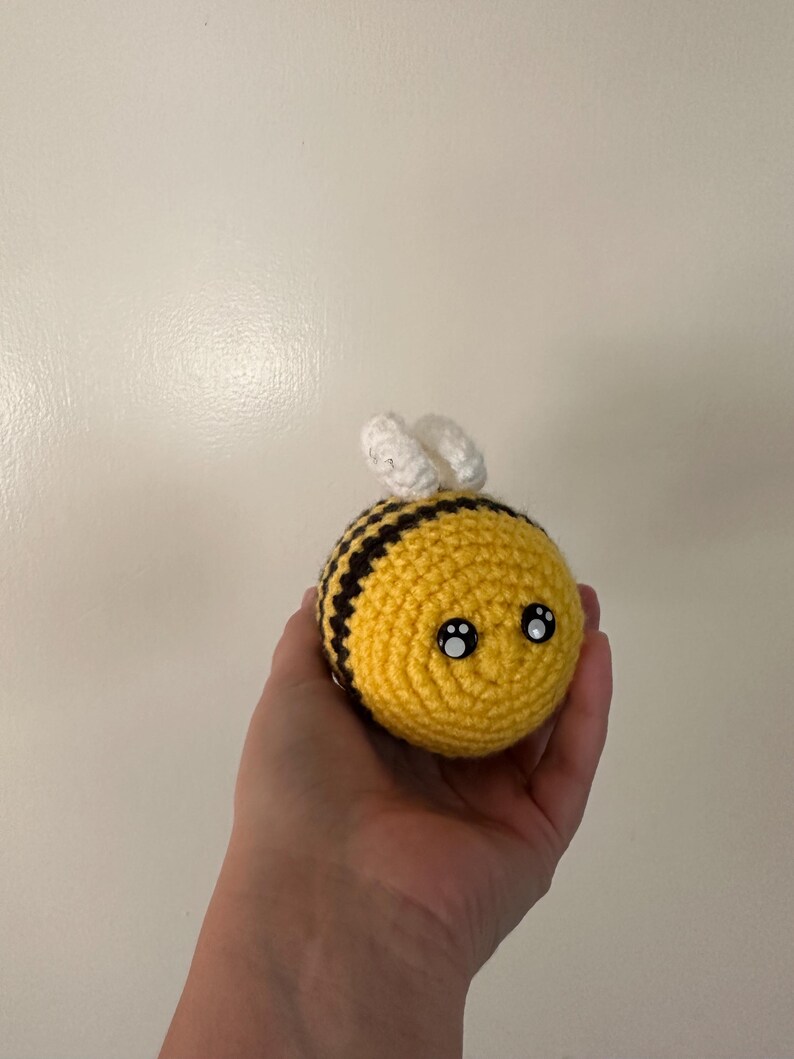 Puede incluir: Una abeja de ganchillo hecha a mano con cuerpo amarillo, rayas negras y alas blancas. La abeja tiene dos ojos negros y blancos, lo que le da una apariencia linda. La abeja se sostiene en una mano.