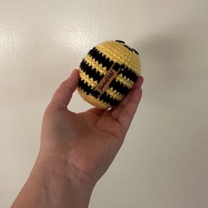Puede incluir: Un juguete de abeja de ganchillo hecho a mano, con rayas amarillas y negras. La abeja tiene una peque&ntilde;a cabeza negra y una etiqueta marr&oacute;n con el texto "JadeePark". El juguete se sostiene en una mano.