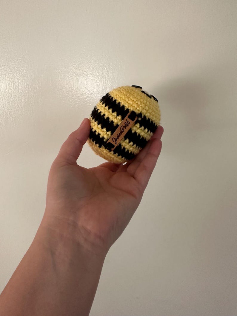 Puede incluir: Un juguete de abeja de ganchillo hecho a mano, con rayas amarillas y negras. La abeja tiene una peque&ntilde;a cabeza negra y una etiqueta marr&oacute;n con el texto "JadeePark". El juguete se sostiene en una mano.
