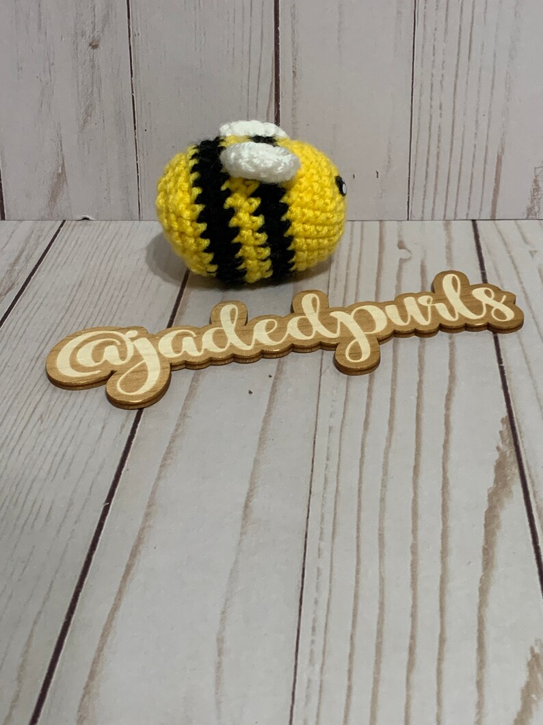 Puede incluir: Una abeja de ganchillo hecha a mano con rayas amarillas y negras, alas blancas y peque&ntilde;os ojos negros. La abeja est&aacute; sobre una superficie de madera. Un letrero de madera con el texto "@jadedpuris" est&aacute; en primer plano.