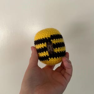Puede incluir: Un art&iacute;culo hecho a mano en forma de abeja, tejido a ganchillo, con rayas amarillas y negras. La abeja tiene una peque&ntilde;a etiqueta marr&oacute;n que dice "Jaded Curls". El art&iacute;culo se sostiene en una mano.