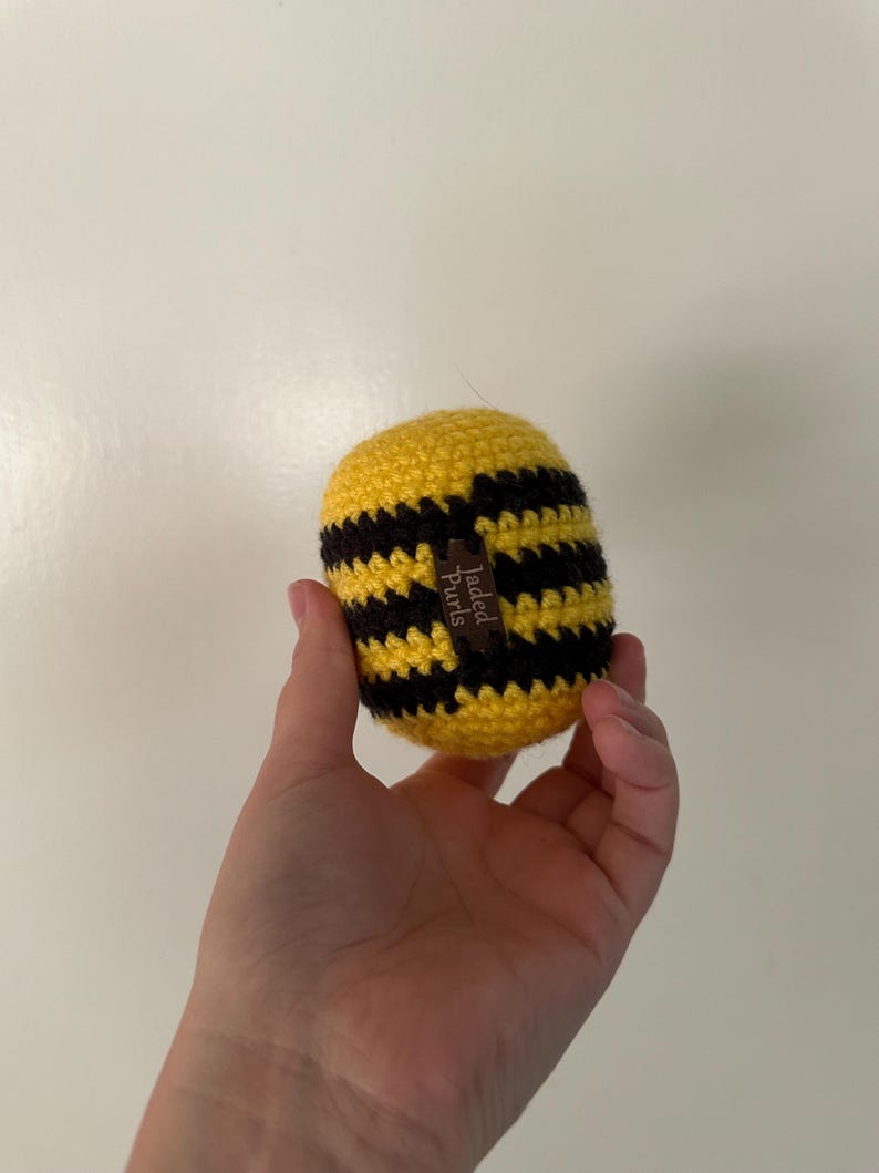 Puede incluir: Un art&iacute;culo hecho a mano en forma de abeja, tejido a ganchillo, con rayas amarillas y negras. La abeja tiene una peque&ntilde;a etiqueta marr&oacute;n que dice "Jaded Curls". El art&iacute;culo se sostiene en una mano.