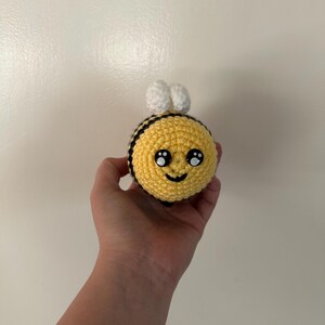 Puede incluir: Un juguete de abeja hecho a mano con ganchillo. La abeja tiene un cuerpo amarillo con rayas negras, alas blancas y una cara sonriente. El juguete se sostiene en una mano sobre un fondo blanco.