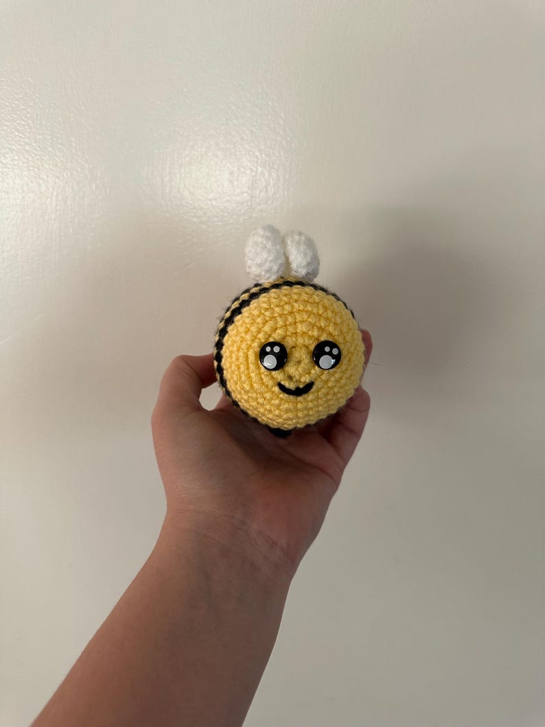 Puede incluir: Un juguete de abeja hecho a mano con ganchillo. La abeja tiene un cuerpo amarillo con rayas negras, alas blancas y una cara sonriente. El juguete se sostiene en una mano sobre un fondo blanco.