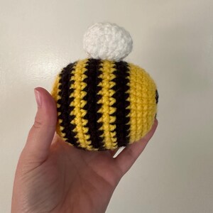 Puede incluir: Una abeja de ganchillo hecha a mano con rayas amarillas y negras y un pomp&oacute;n blanco en la parte superior. La abeja se sostiene en una mano sobre un fondo liso. Las puntadas de ganchillo son visibles, dando a la abeja una apariencia texturizada.