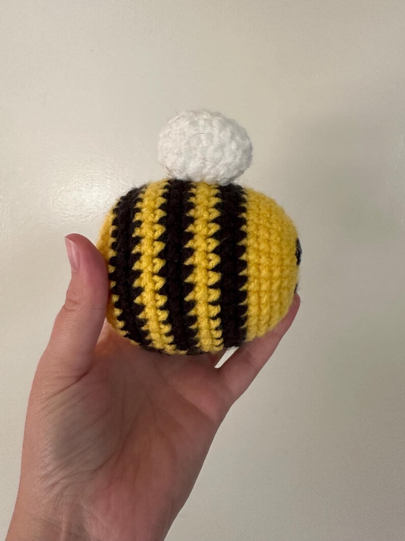 Puede incluir: Una abeja de ganchillo hecha a mano con rayas amarillas y negras y un pomp&oacute;n blanco en la parte superior. La abeja se sostiene en una mano sobre un fondo liso. Las puntadas de ganchillo son visibles, dando a la abeja una apariencia texturizada.
