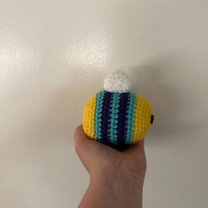 Puede incluir: Un juguete de abeja hecho a mano con ganchillo. La abeja presenta rayas amarillas, turquesas y moradas, con un pomp&oacute;n blanco en la parte superior. Se utiliza una peque&ntilde;a cuenta negra para el ojo. El juguete se sostiene en una mano.