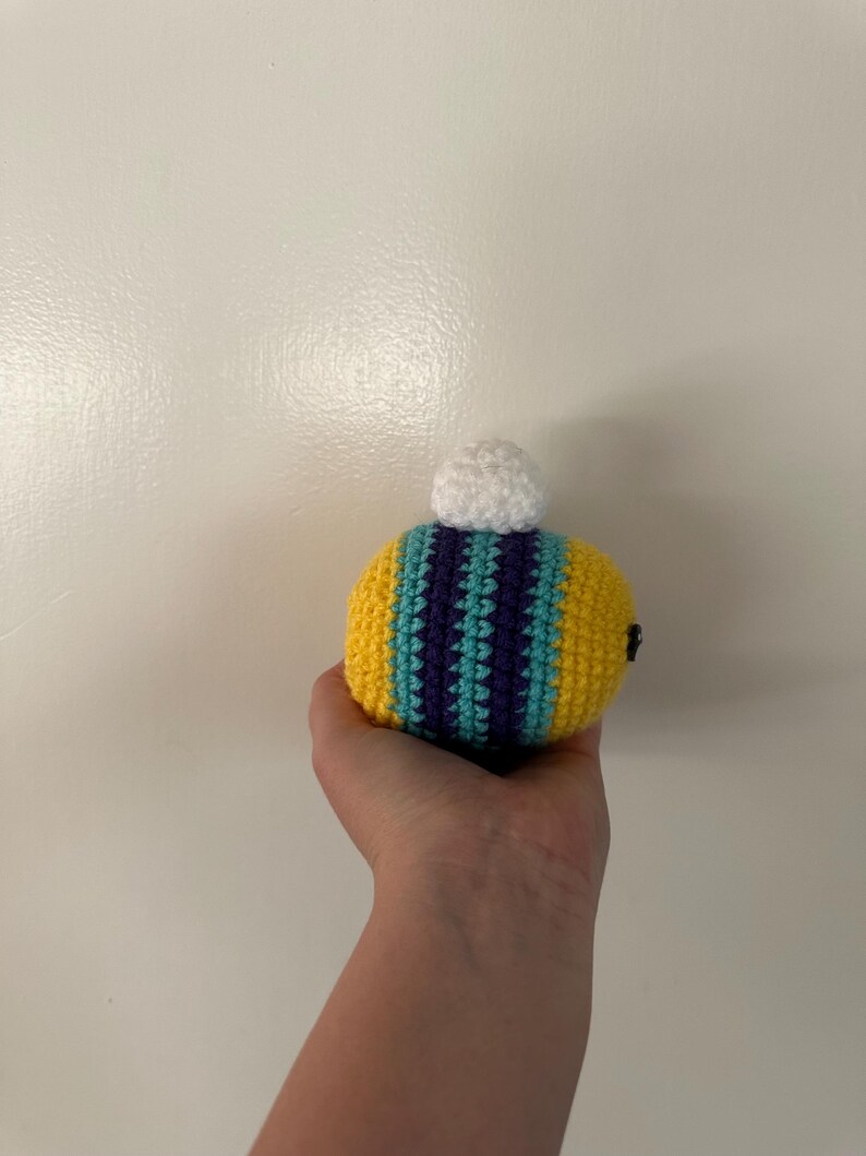 Puede incluir: Un juguete de abeja hecho a mano con ganchillo. La abeja presenta rayas amarillas, turquesas y moradas, con un pomp&oacute;n blanco en la parte superior. Se utiliza una peque&ntilde;a cuenta negra para el ojo. El juguete se sostiene en una mano.
