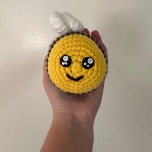 Puede incluir: Un juguete de abeja hecho a mano, tejido a ganchillo, con cuerpo amarillo, rayas negras y alas blancas. La abeja tiene una cara sonriente con ojos grandes. El juguete es redondo y suave.