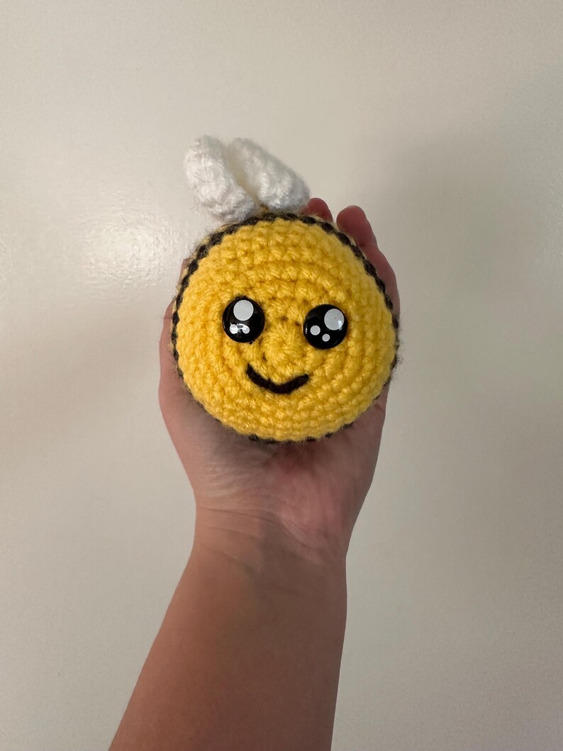 Puede incluir: Un juguete de abeja hecho a mano, tejido a ganchillo, con cuerpo amarillo, rayas negras y alas blancas. La abeja tiene una cara sonriente con ojos grandes. El juguete es redondo y suave.