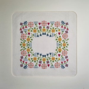 以下が含まれることがあります： ピンク、黄色、青、緑など、さまざまな色の花のボーダーが特徴の正方形のクロスステッチデザイン。デザインには花、ハート、蝶が含まれており、中央には空白の白い正方形があります。