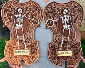 May include: Two wooden violin-shaped plaques with intricate carvings. Each plaque features a skeleton holding a ring, surrounded by ornate floral designs. The text "YOUR NAME" is engraved on each plaque.