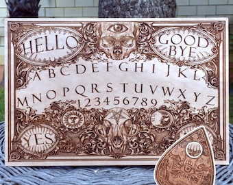 May include: A wooden Ouija board with an intricate design featuring a skull, an eye, and a pentagram. The board includes the alphabet, numbers, "yes", "no", "hello", and "goodbye". A wooden planchette with an eye design is also included.
