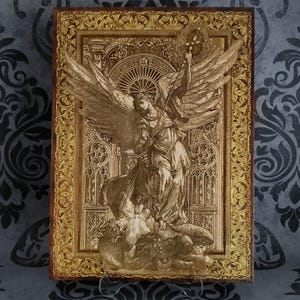 Könnte beinhalten: Eine rechteckige Holztafel mit einer lasergravierten Darstellung eines Engels mit großen Flügeln, der über einer gefallenen Figur steht. Das zentrale Bild ist von einem verzierten, goldfarbenen Rand mit komplexen Blumenmustern umgeben.