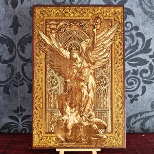 Op de afbeelding: Een rechthoekige houten plaat met een lasergegraveerde afbeelding van een engel met grote vleugels die boven een gevallen figuur staat. Het kunstwerk is omlijst door een versierde, goudkleurige rand met complexe gotische architecturale details.