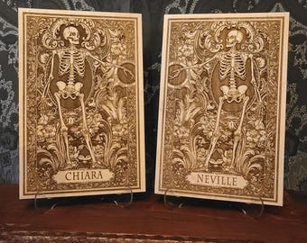 Puede incluir: Dos placas de madera con diseños de esqueletos grabados. Cada placa presenta una figura de esqueleto detallada rodeada de patrones florales. Los nombres "Chiara" y "Neville" están inscritos en la parte inferior de cada placa. Las placas se exhiben sobre soportes metálicos.