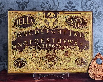 May include: A rectangular wooden spirit board featuring intricate gold-toned engravings of a skull, an eye, a sun, a moon, and ornate scrollwork. The board displays the alphabet, numbers zero through nine, and the words HELLO, GOOD BYE, YES, and NO.