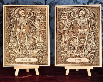 May include: Two rectangular wooden plaques featuring laser-engraved skeleton figures surrounded by intricate floral and scrollwork patterns. The left plaque displays the name AMBER, and the right plaque displays the name BENJ. Each rests on a small easel.