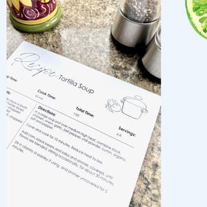 K&ouml;nnte beinhalten: Ein gedrucktes Rezept f&uuml;r Tortilla-Suppe auf einem wei&szlig;en Blatt Papier. Das Rezept enth&auml;lt Kochzeit, Gesamtzeit und Anweisungen. Eine Zeichnung eines Topfes ist enthalten. Das Rezept liegt auf einer Arbeitsplatte mit Salz- und Pfefferstreuer.