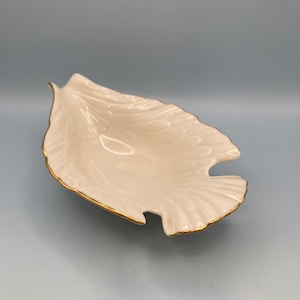 May include: A cream-colored, leaf-shaped dish with a gold trim. The dish has a textured surface and a unique cutout design. The elegant serving dish is perfect for displaying small items or serving snacks. The dish is made of porcelain.