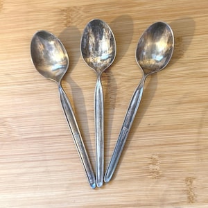 May include: Three vintage silver spoons arranged on a wooden surface. The spoons have oval bowls and slender handles. The silver has a tarnished appearance, suggesting age and use. The spoons are likely for serving or eating.