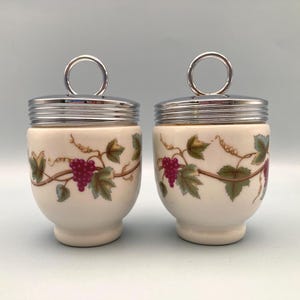 May include: A pair of off-white ceramic egg coddlers featuring a painted pattern of purple grape clusters and green leaves. Each coddler has a polished silver-tone metal lid with a circular ring handle on top for easy lifting and opening.