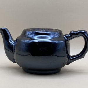 May include: A glossy black ceramic teapot with a curved spout and a decorative handle. The teapot has a rounded body and a lid with a small knob. The surface reflects light, highlighting its smooth finish.