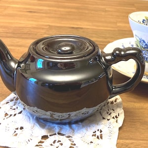 May include: A glossy black ceramic teapot with a rounded body, a curved spout, and a matching lid. Beside the teapot sits a white porcelain teacup and saucer decorated with a blue floral pattern, all resting on a white lace doily.