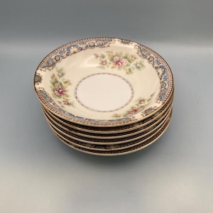 May include: A stack of six cream-colored porcelain bowls. Each bowl features a floral design with pink and blue flowers, a blue and gold border, and a gold rim. The bowls are stacked on a light blue surface.