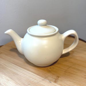 May include: A solid off-white ceramic teapot with a rounded body, a curved spout, a matching lid with a small knob handle, and a side handle. The teapot features a smooth, matte finish and a simple, functional design for brewing and serving tea.