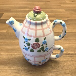 May include: A ceramic tea-for-one set featuring a teapot stacked on a teacup. The white surface is decorated with a pink plaid pattern, blue flowers, green leaves, and a central rose. The lid is light green with white polka dots and a pink rose knob.