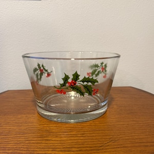 May include: A clear glass bowl with a festive holly and berry design. The bowl features green holly leaves and red berries, perfect for holiday decor. The bowl has a wide, flared shape and a textured base.