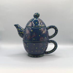 May include: A ceramic tea-for-one set featuring a dark blue teapot stacked on a matching teacup. The set is decorated with small lime green polka dots and dark reddish-brown floral patterns. The teapot has a lid with a small knob handle.