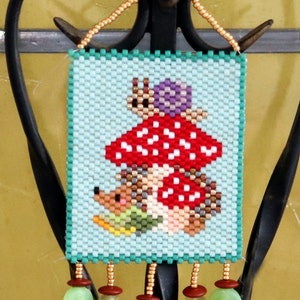 Op de afbeelding: Een kralenwanddecoratie met een blauwe achtergrond waarop een rode en witte paddenstoel met een slak erop te zien is. Twee egels bevinden zich onder de paddenstoel. De decoratie heeft groene, blaadjesvormige kralen.