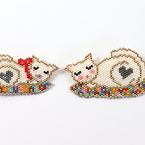 May include: Two white beaded cat brooches with gray hearts on their backs, lying on colorful beaded flower beds.