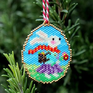 May include: A beaded bauble featuring a white bunny with a red scarf, holding a basket of colourful eggs. The bunny is on a green and purple background with a blue sky.